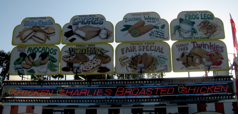 Signs above a food booth offering all kinds of 'Totally Fried' foods that you wouldn't normally fry, like pizza, avocados, Twinkies and s'mores.