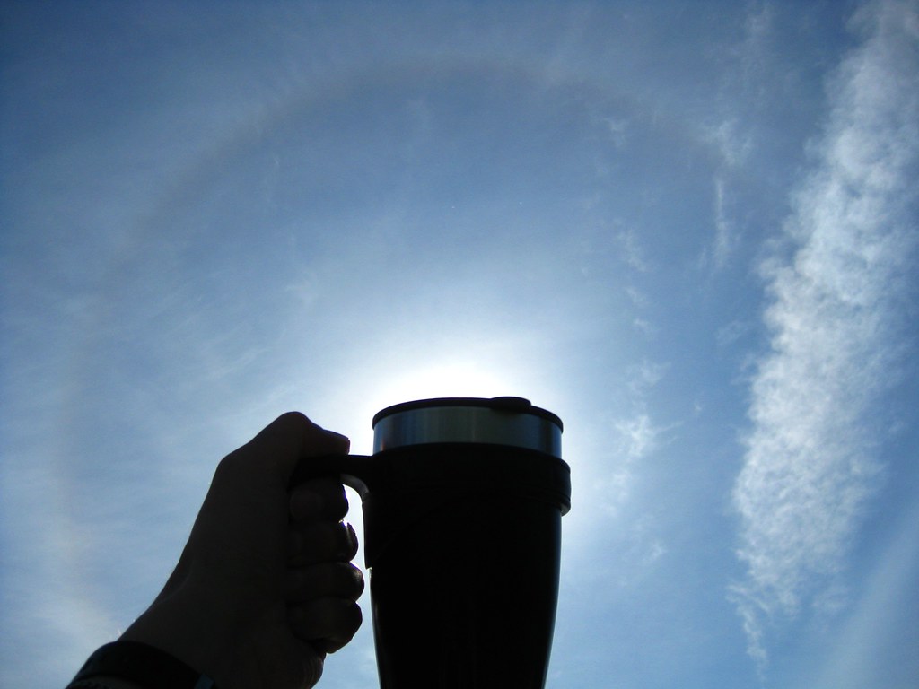 A bright ring surrounds the sun, which is blocked by the silhouette of a hand holding up a coffee mug.
