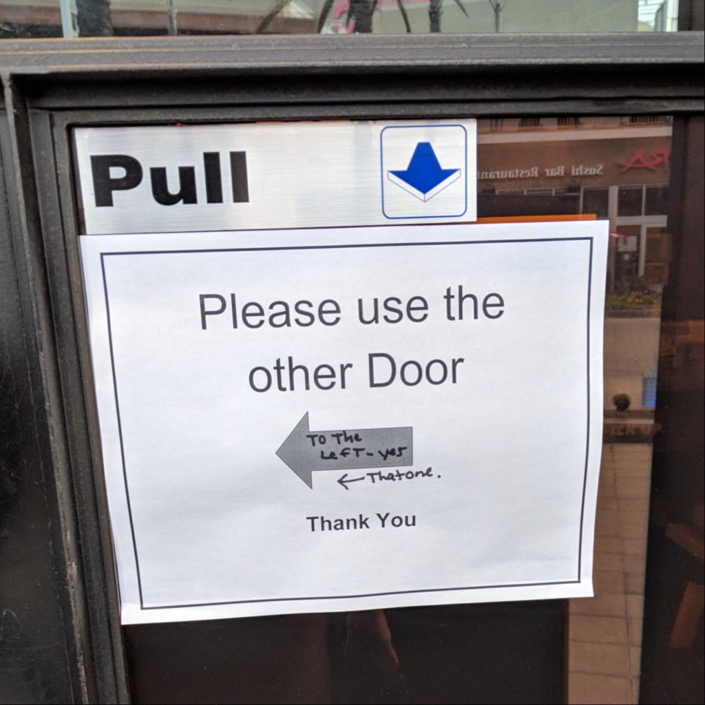Printed sign: Please use the other Door (with arrow). Hand written on it: To the left - yes, that one (with another arrow)