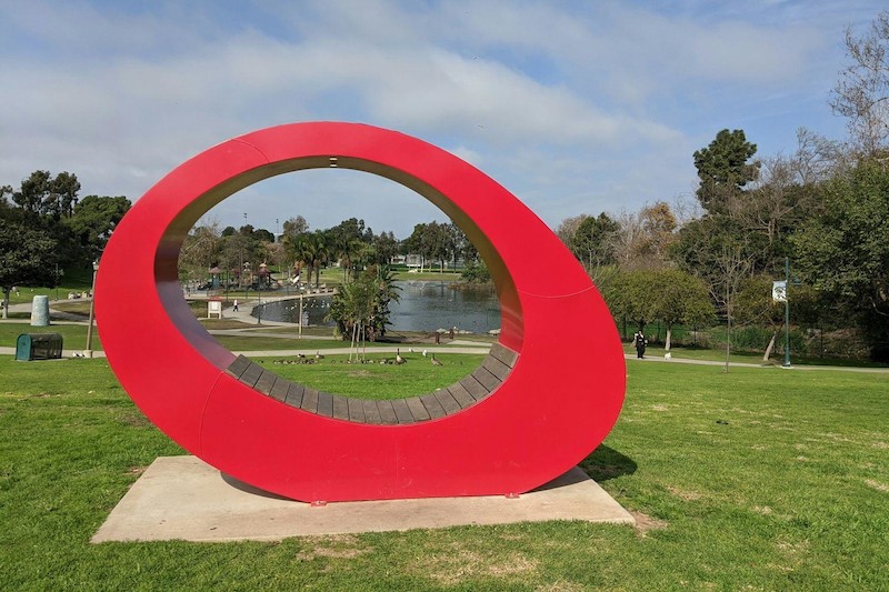A red irregular ring sculpture, big enough to climb through, in a city park on a sunny day.