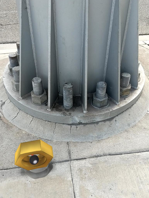A large metal post held to the ground by *huge* bolts. One is missing a nut. Nearby, a Meltan (a Pokemon whose head is a giant hex nut) appears to be moving away from the post.