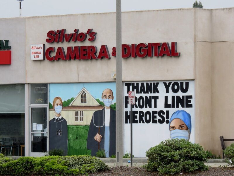 Camera shop with windows painted with American Gothic with six feet of social distancing and face masks, and a thank you message to front line heroes in scrubs.