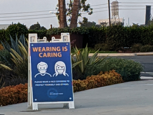 Free-standing temporary sign on a sidewalk corner proclaiming WEARING IS CARING and showing two cartoon people, one with short hair and the other with long hair, wearing face masks covering their mouths and nose.