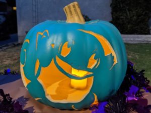 An artificial teal pumpkin carved with an image od a Squirtle, a turtle-like Pokemon that is typically light blue, lit from inside.